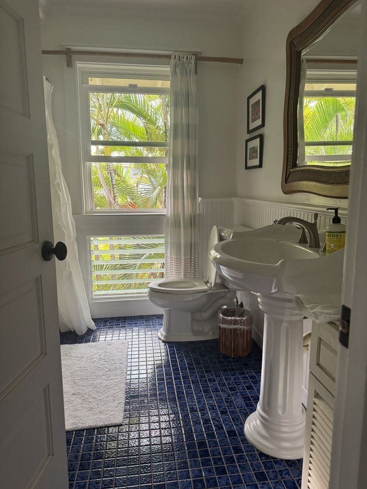 A sparkling clean bathroom with a pedestal sink, blue tile floor and palm trees outside the window